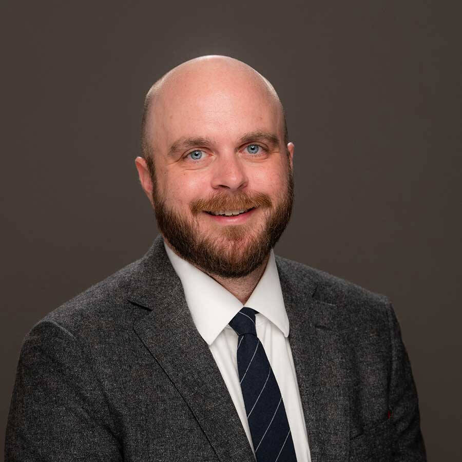 Justin Melick sporting a dark grey suit with a navy blue tie and a white shirt